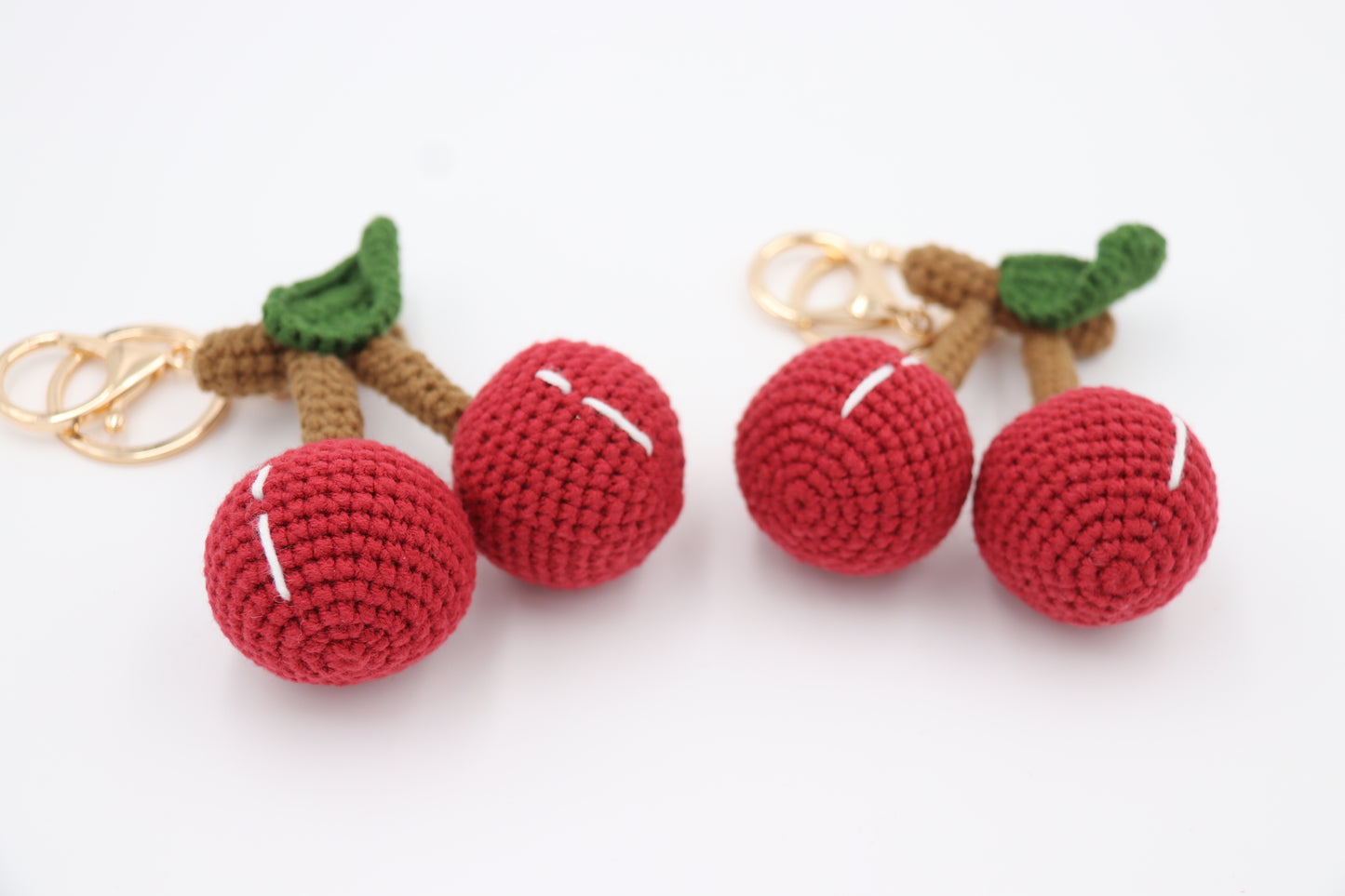 Two hand-crochet cherry keychains with red fruit, brown stems, green leaves, and gold metal rings on a white background.