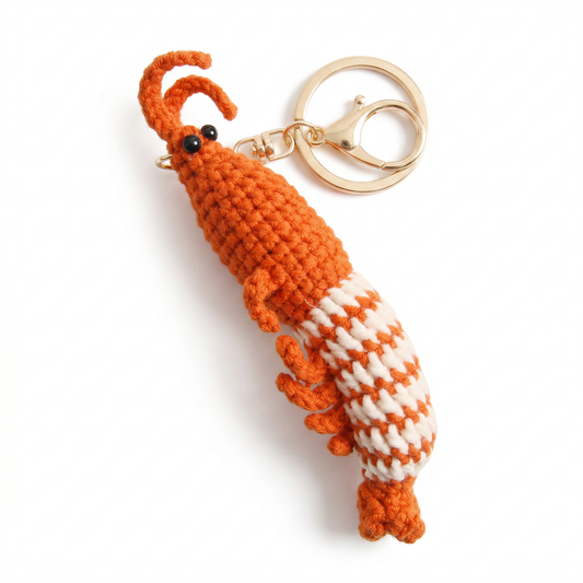 A crochet prawn keychain featuring orange and white wool patterns held by a hand against a white background.